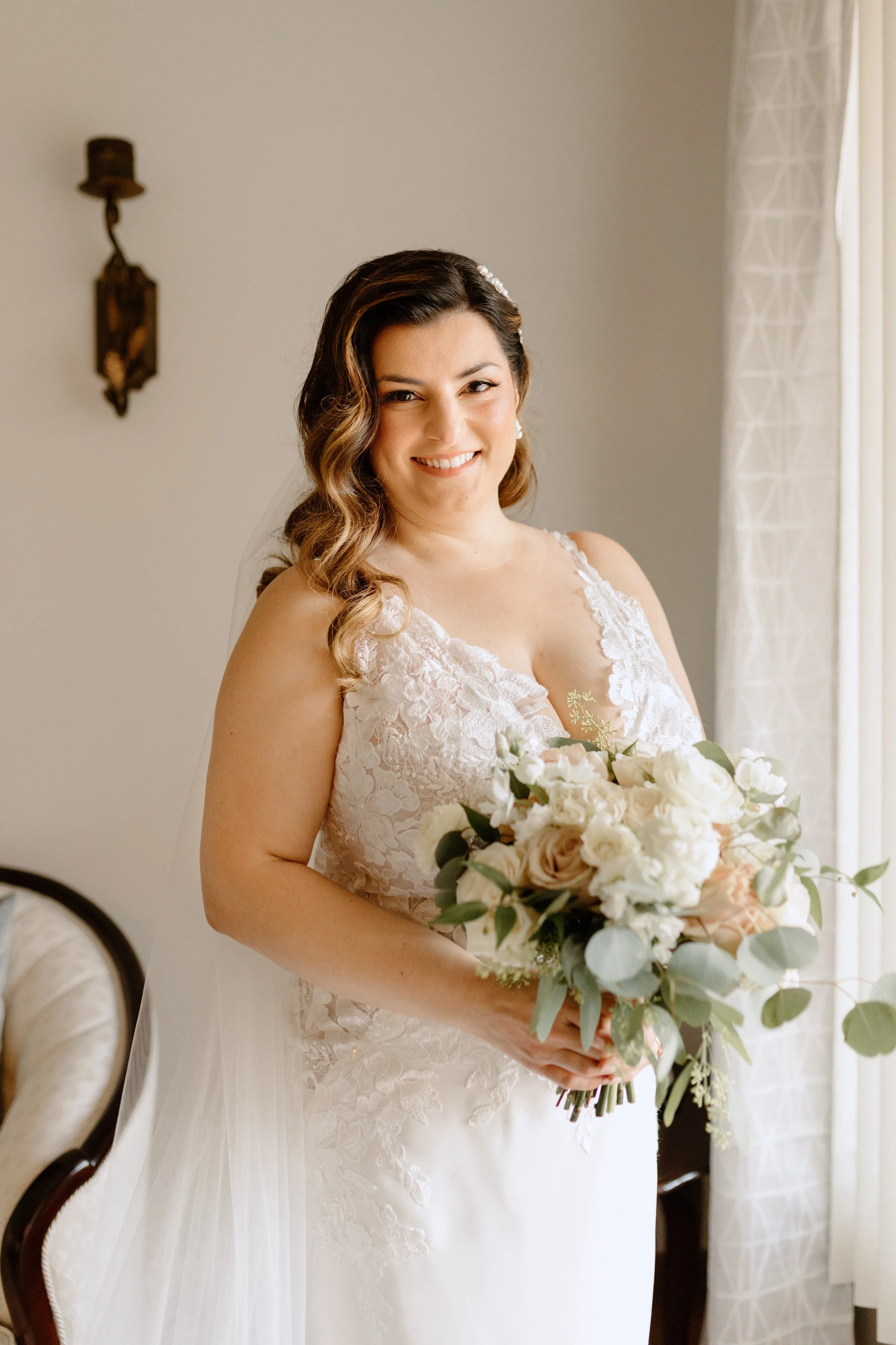 Bride in white lace wedding dress holding bouquet, smiling indoors.