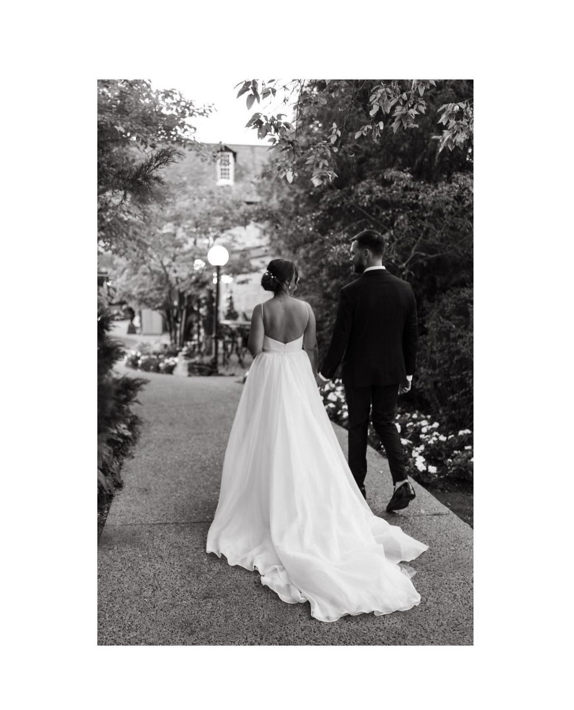 Bride and groom walking on a garden path, bride in a white dress and groom in a suit, black and white photo.