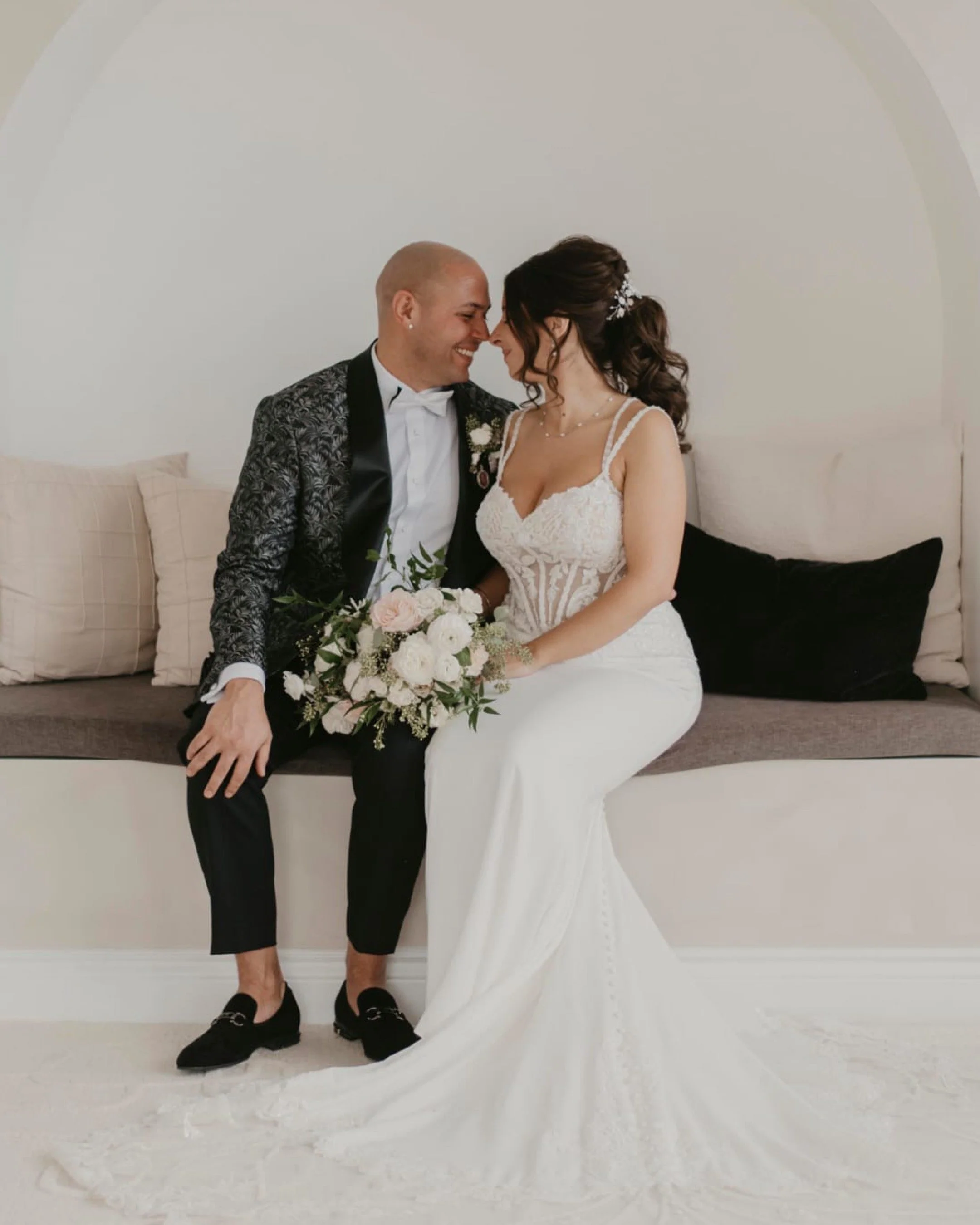 Bride and groom sitting closely on a bench, bride in a white gown holding a bouquet, groom in a patterned jacket and black pants, both smiling fondly.