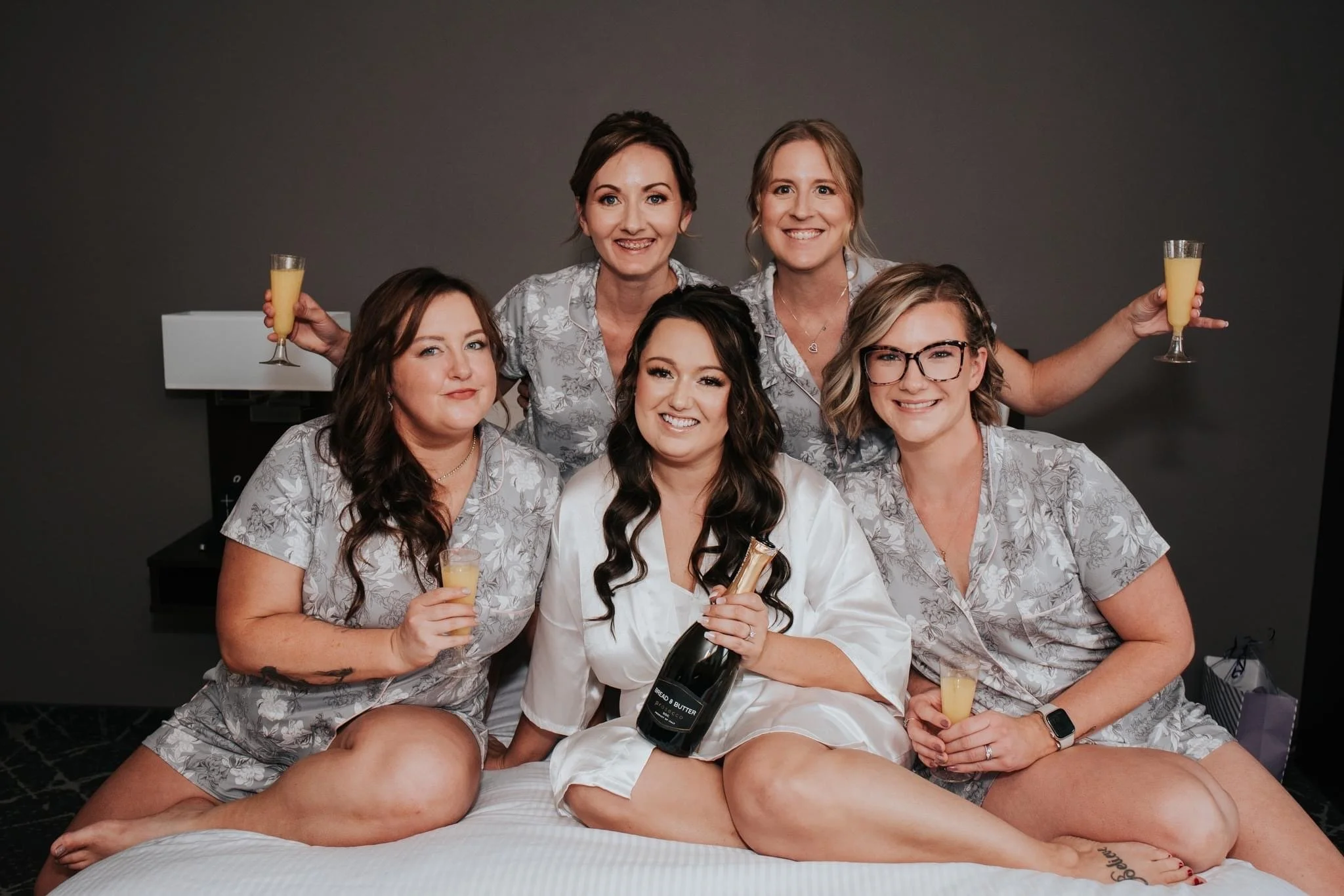 A group of women sitting on a bed, each holding a glass of orange juice, with one woman in a white robe holding a bottle of wine.