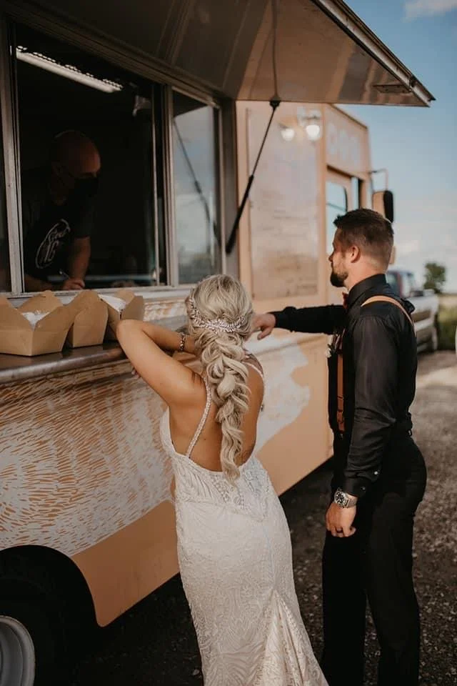 A bride and groom ordering food from a food truck, with takeout boxes on the counter.