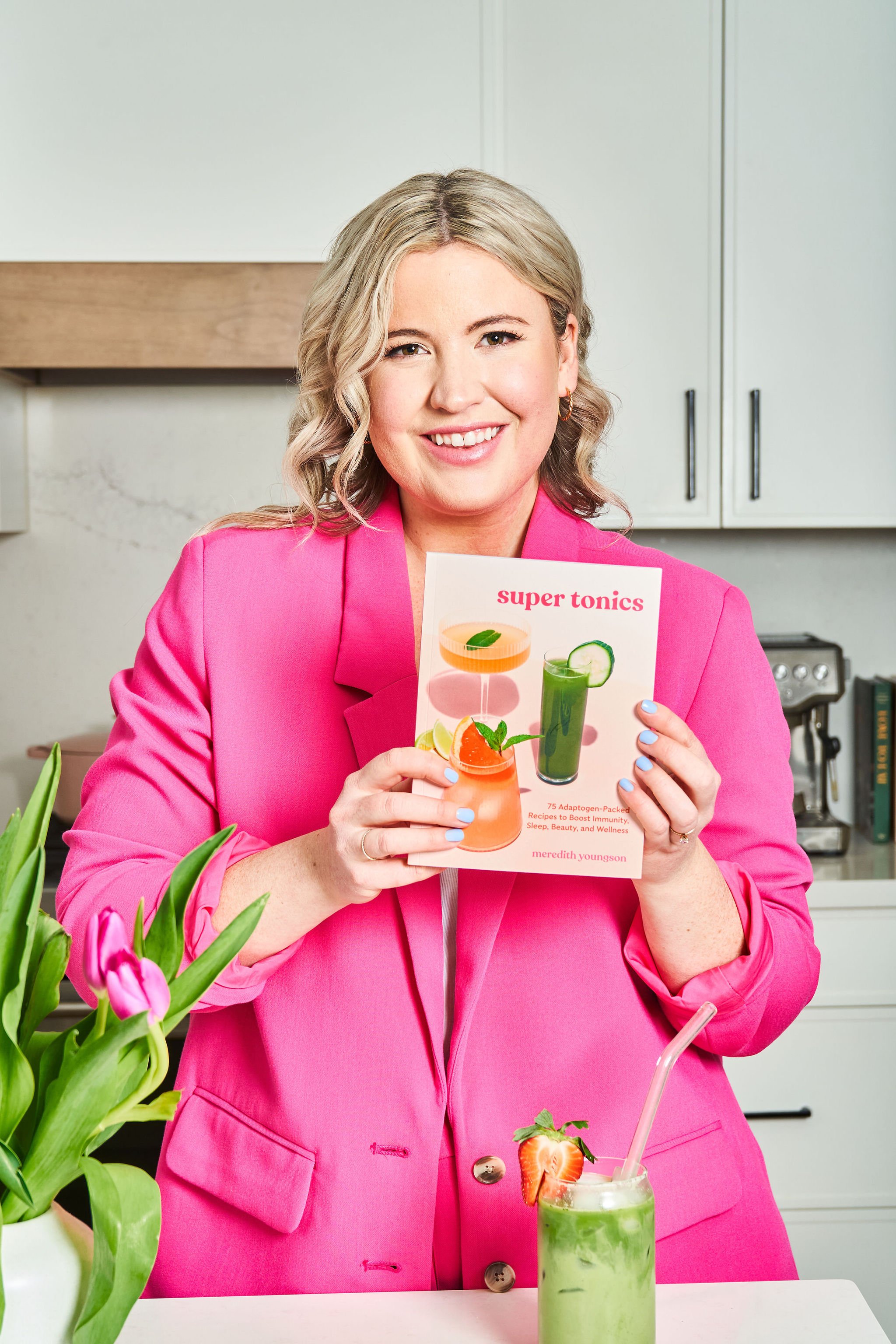 Person in a pink jacket holding a book titled "Super Tonics" in a kitchen with a green drink garnished with a strawberry.