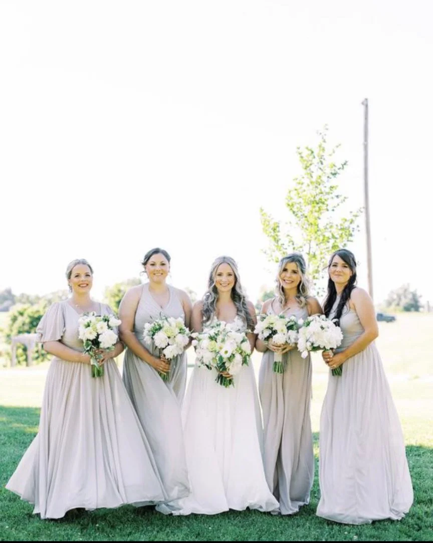 Five women in light gray and white gowns holding bouquets standing on grass outdoors, possibly bridesmaids and a bride.