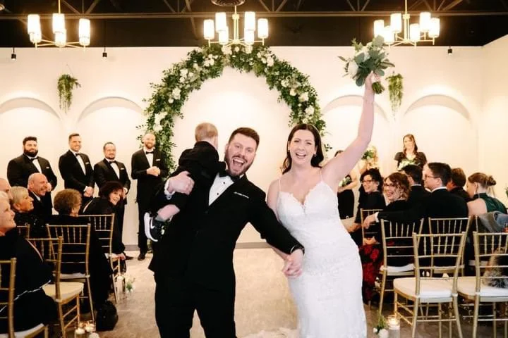 Bride and groom celebrating with guests at wedding ceremony, holding a child, greenery arch behind.