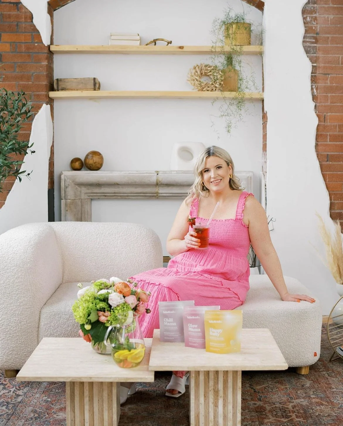 Woman in a pink dress sitting on a cream sofa holding a drink, with a coffee table in front displaying flower arrangements and three colorful wellness product packages.