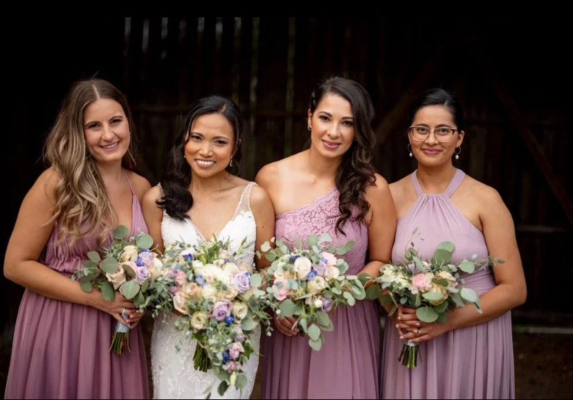 Bride with bridesmaids holding bouquets, wearing lavender dresses indoors.