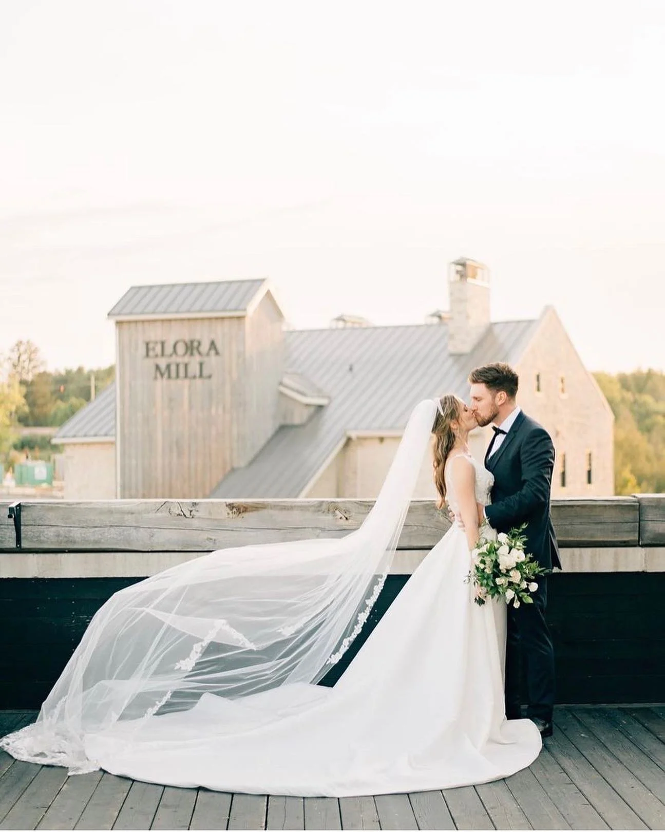 Bride and groom kissing at Elora Mill with bride in white gown and veil, holding bouquet.