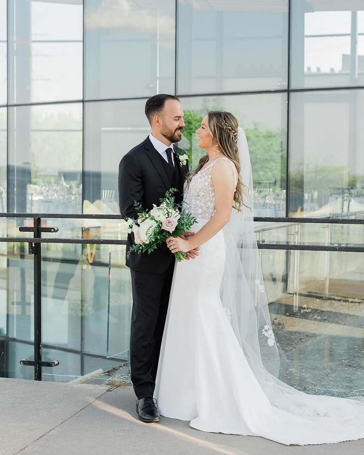 Bride and groom smiling at each other, bride holding bouquet in front of large glass building.