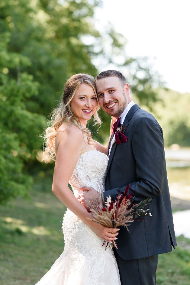 Bride and groom posing outdoors in wedding attire, with the bride holding a bouquet, surrounded by greenery.