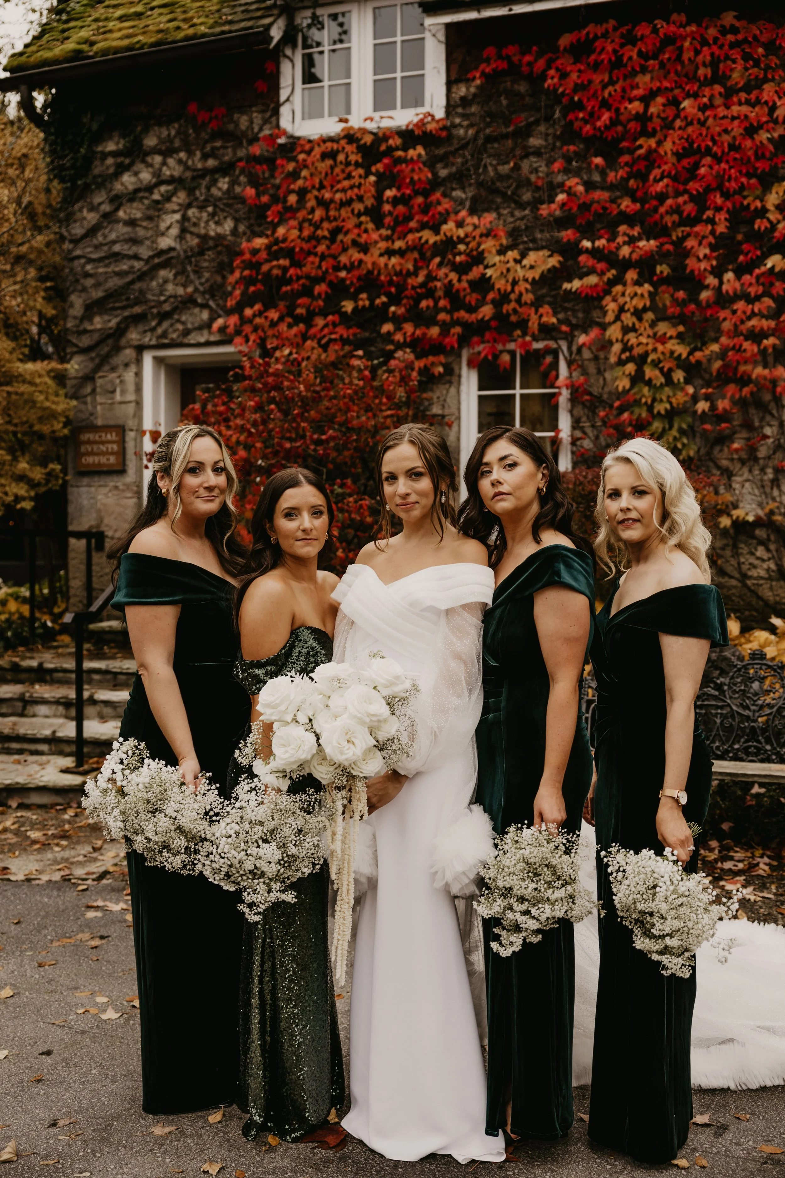 Bride with bridesmaids in green dresses holding bouquets, standing in front of a stone building with red vine foliage.