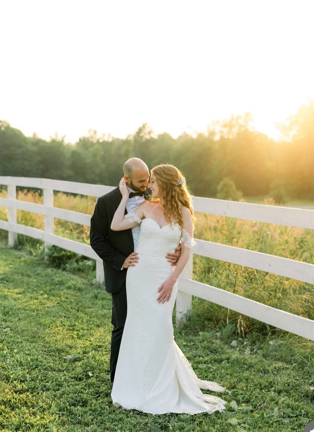 Bride and groom embracing in a field near a white fence, at sunset.