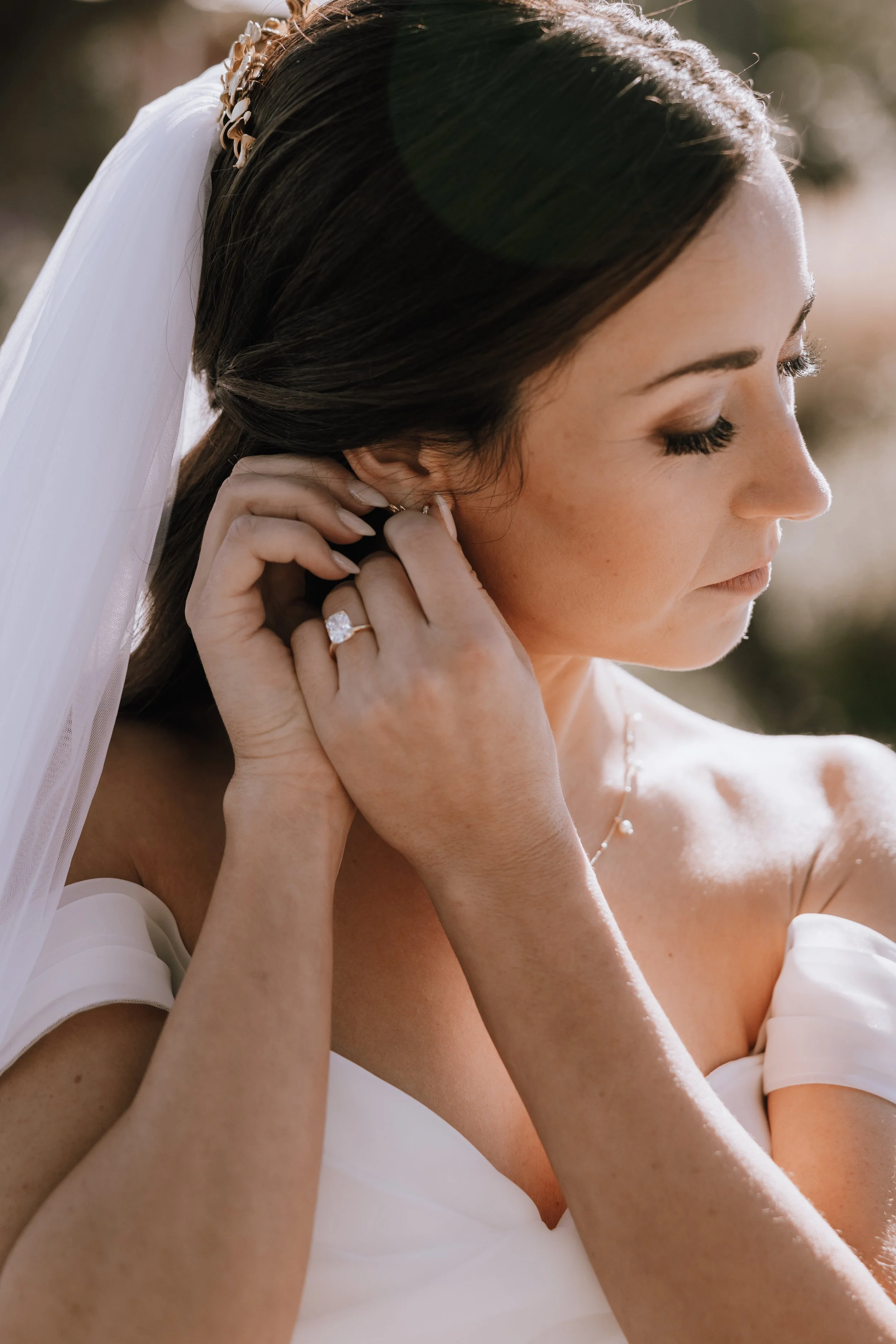 A bride with dark hair and light skin, wearing a white off-shoulder wedding dress and a veil, puts on her earring outdoors in sunlight.
