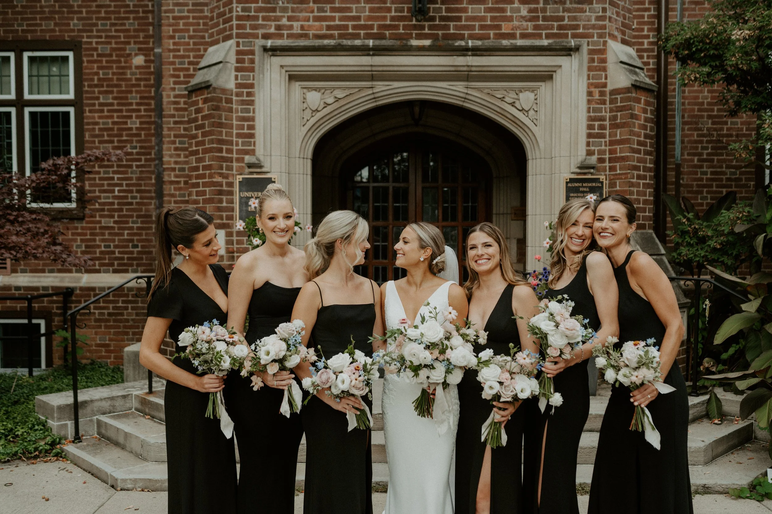 Bride with bridesmaids in black dresses holding bouquets in front of a brick building.