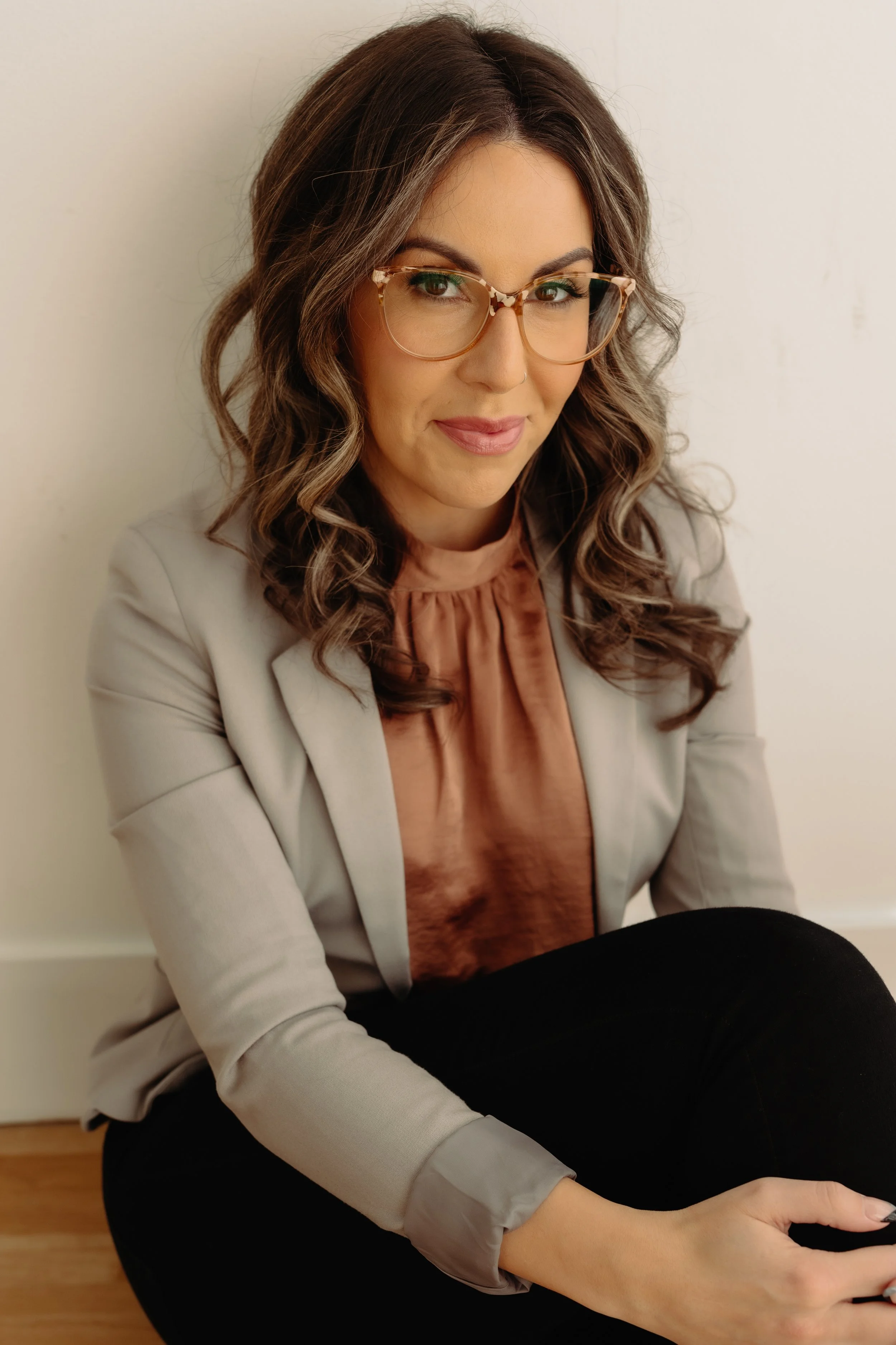 Woman with wavy brown hair, wearing glasses, a beige blazer, and a brown blouse, sitting against a white wall.