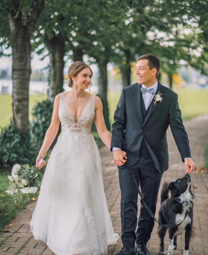 Bride and groom walking with a dog on a pathway, surrounded by trees.