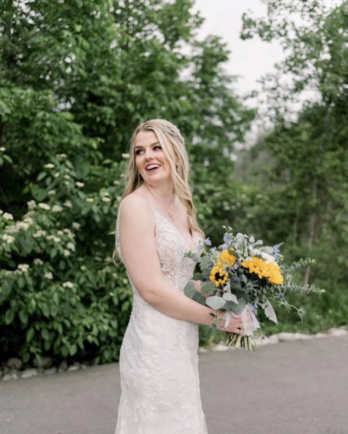 Bride in lace wedding dress holding a bouquet with yellow and blue flowers, standing outdoors with greenery in the background.