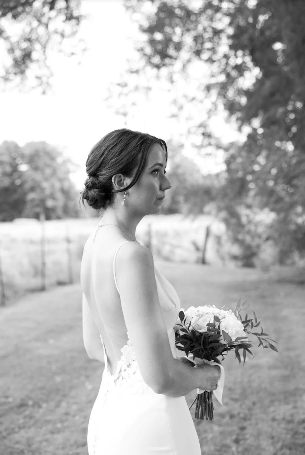 Black and white photo of a bride in a wedding dress holding a bouquet, standing outdoors.
