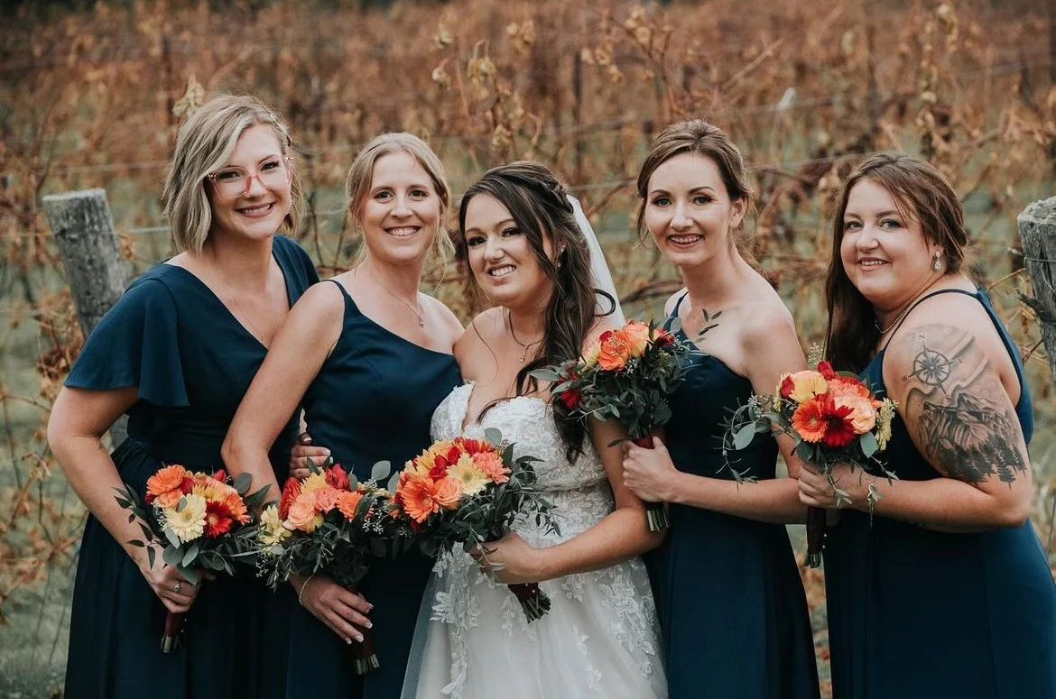 Bride with bridesmaids in navy dresses holding bouquets in a vineyard setting.