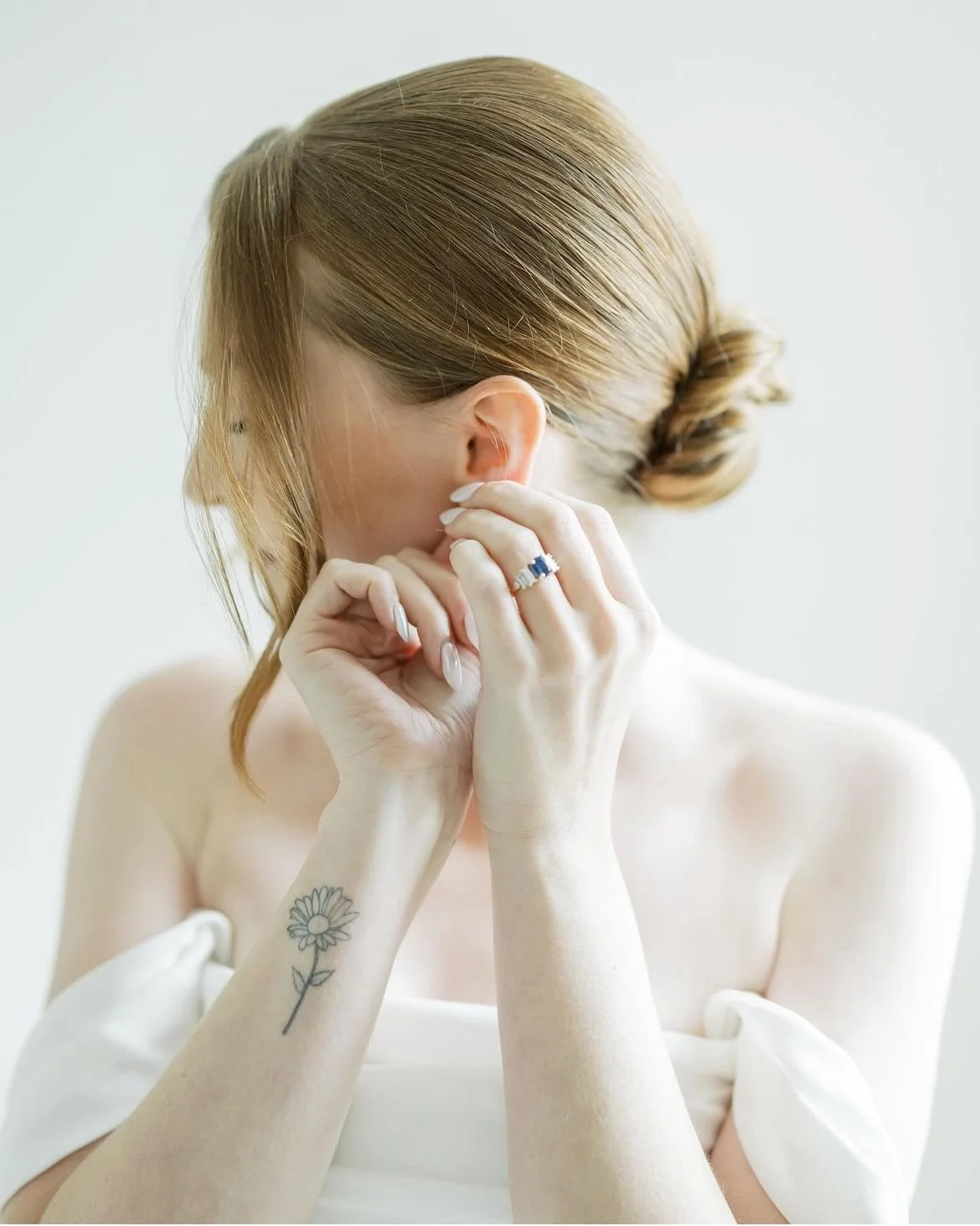 Woman with brown hair in a bun adjusting earrings, wearing a ring, and revealing a daisy tattoo on her forearm, in a white off-shoulder dress.