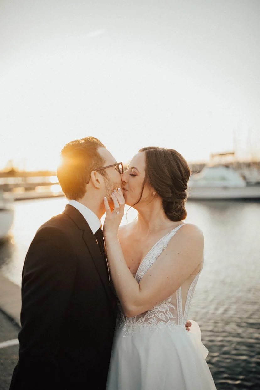 Couple kissing near water with sunset in background, bride in a wedding dress, groom in a suit.