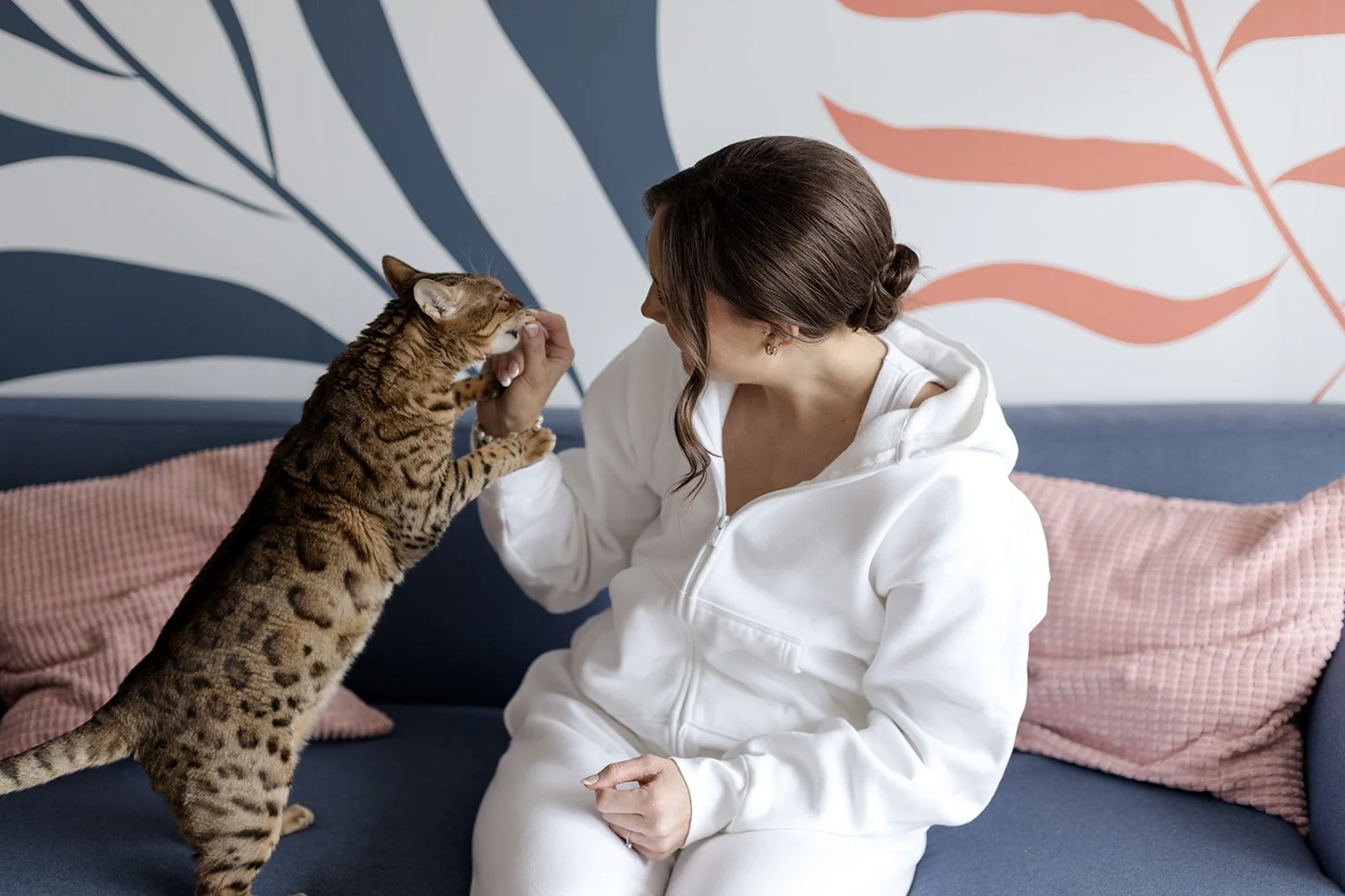 A woman in a white hoodie sitting on a navy blue couch, holding a toy and playing with a Bengal cat, against a background of a wall with blue and pink leaf patterns and pink cushions.