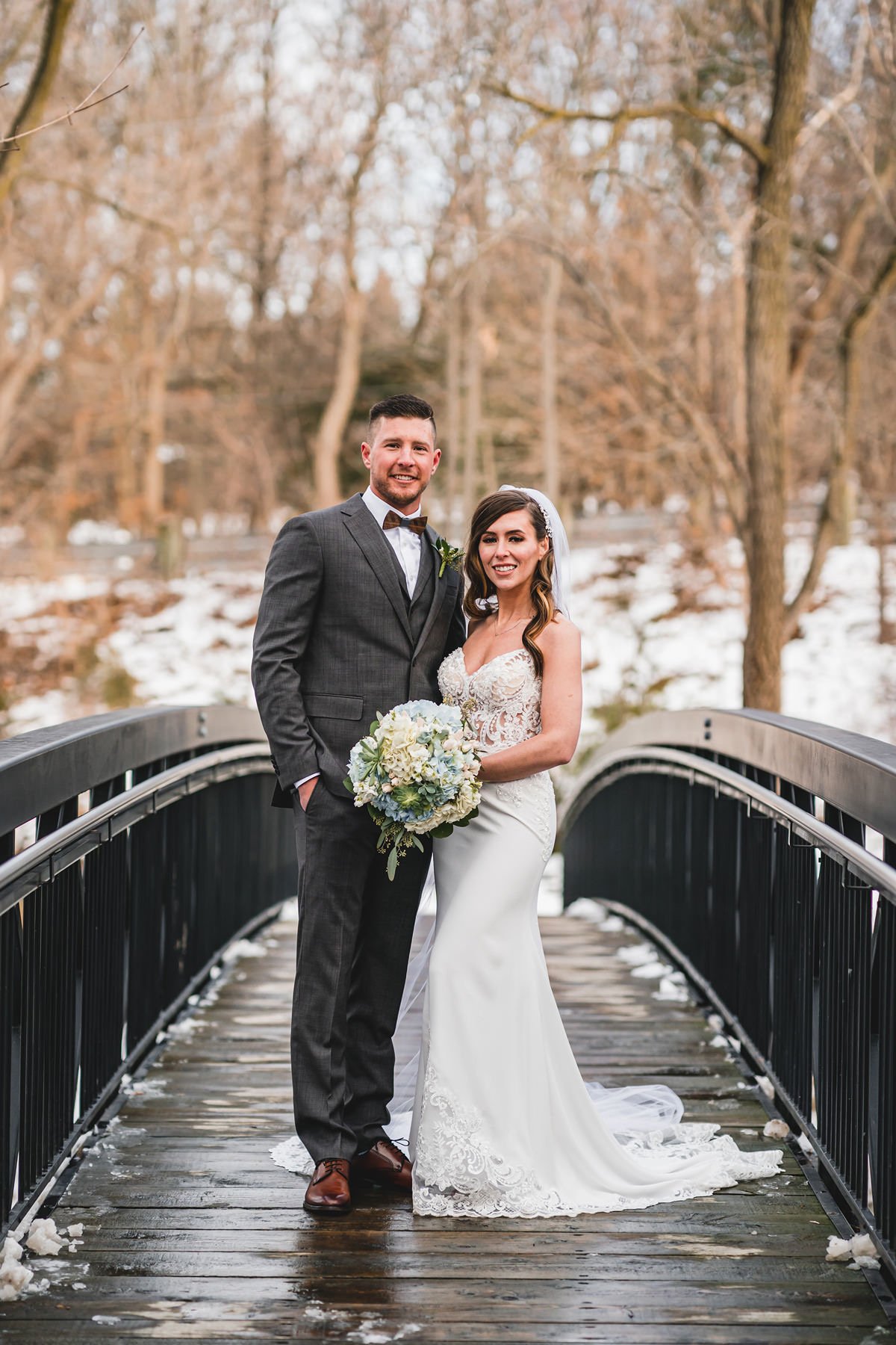 A bride and groom standing on a wooden bridge in a snowy forest, with the bride holding a bouquet of flowers.