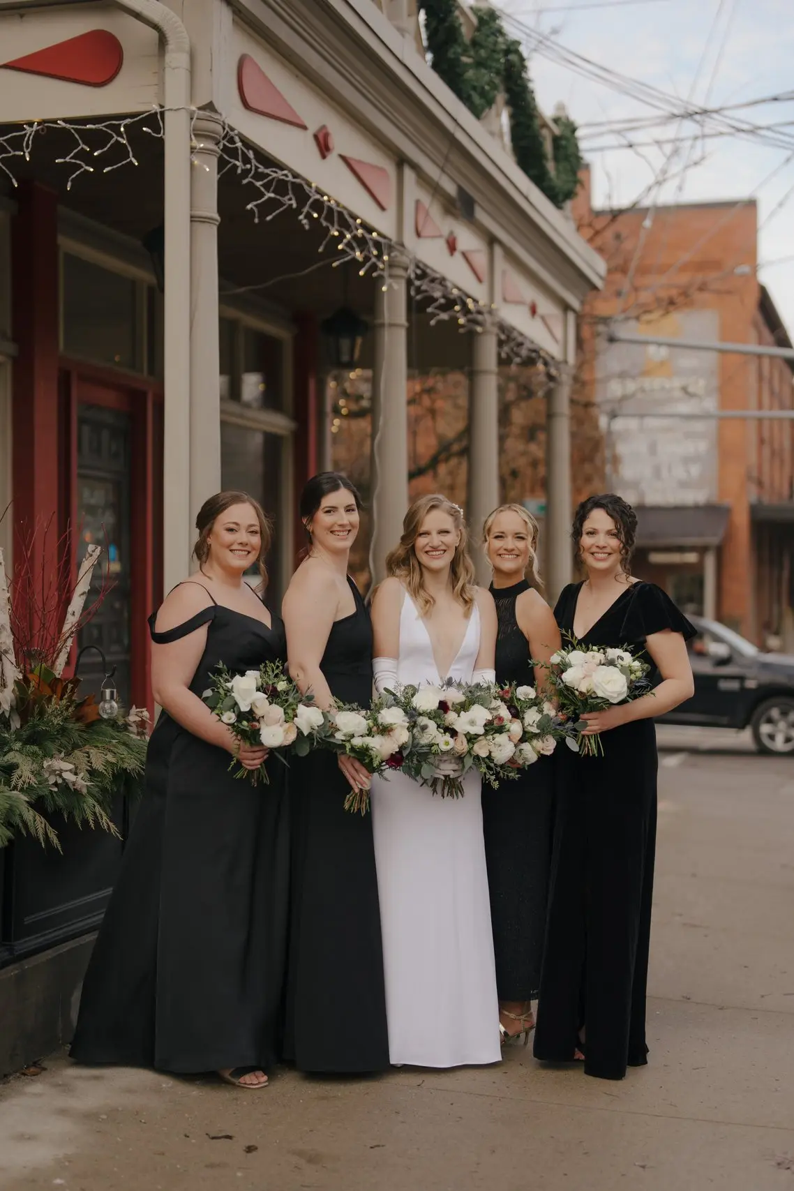 Five women in elegant dresses, one in white and the others in black, holding bouquets, posing outside a building with holiday decorations.