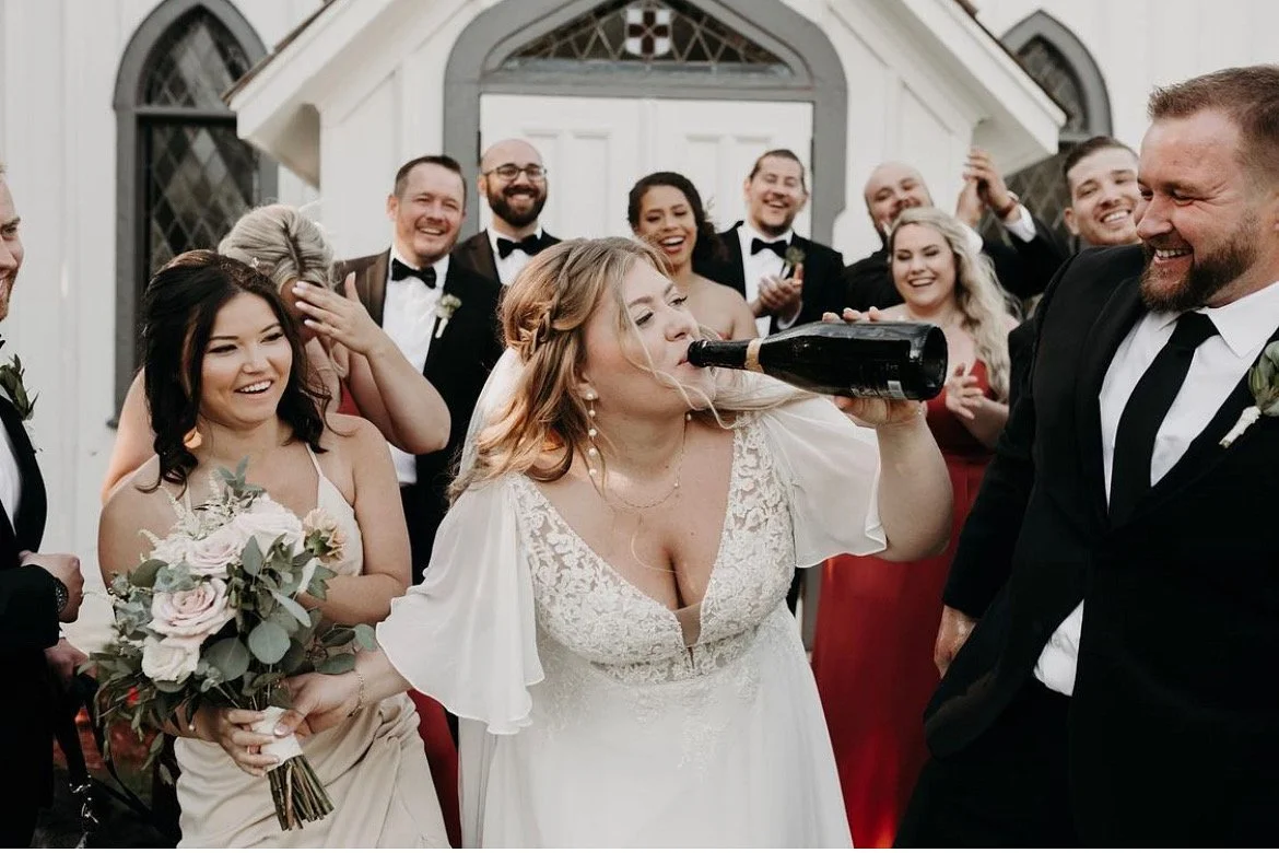 Bride drinking from champagne bottle with wedding party laughing, posing outside a church.