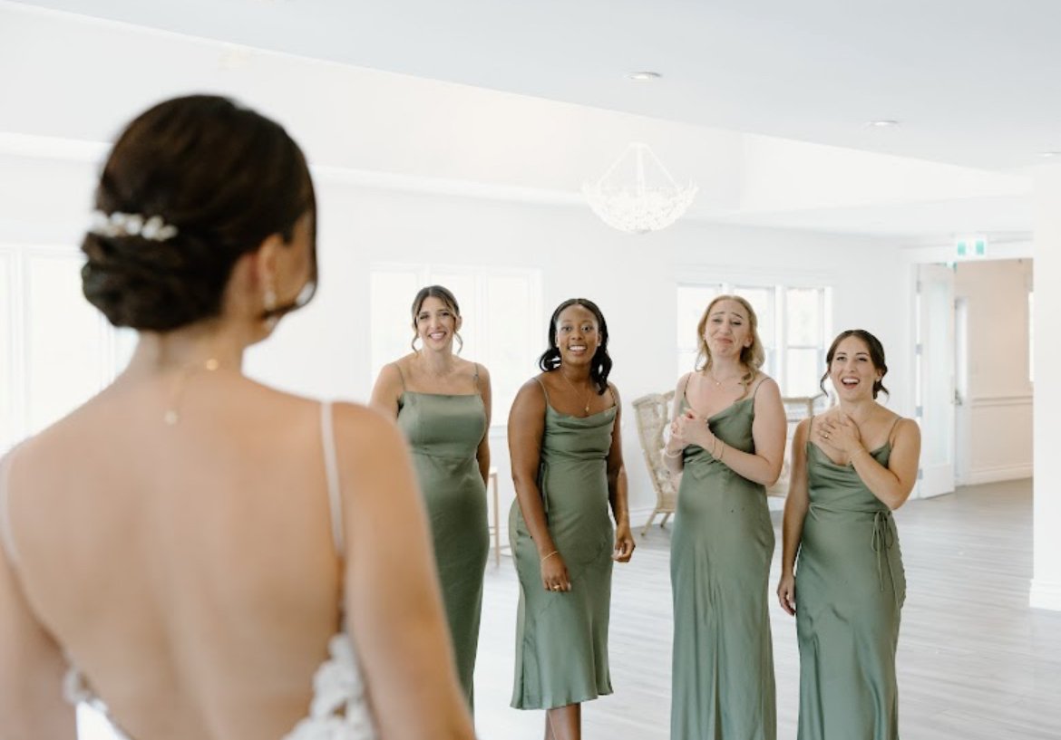 Bride standing with her back to the camera, facing four bridesmaids in green dresses who are smiling and reacting emotionally indoors.