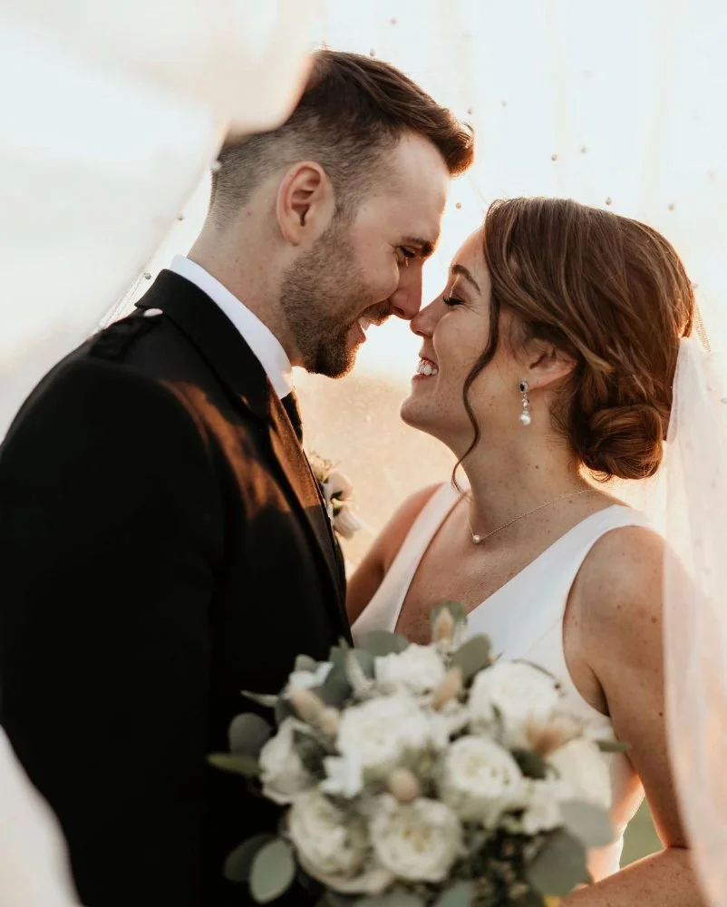 Bride and groom smiling, holding a bouquet, under a veil.