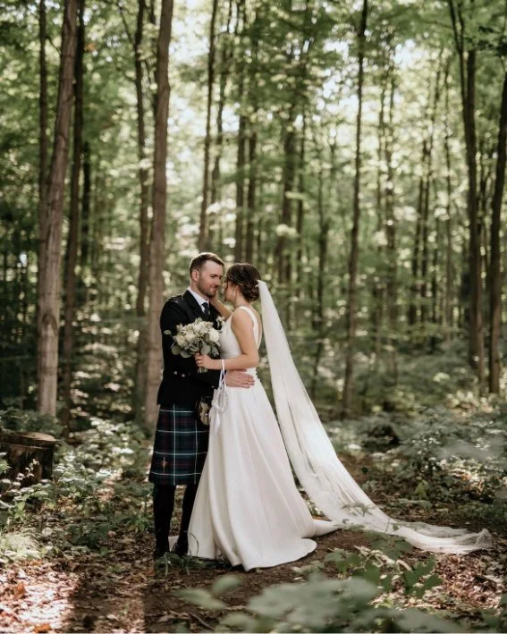Bride and groom embracing in a forest setting, groom in kilt, bride in white dress with long veil, holding bouquet.