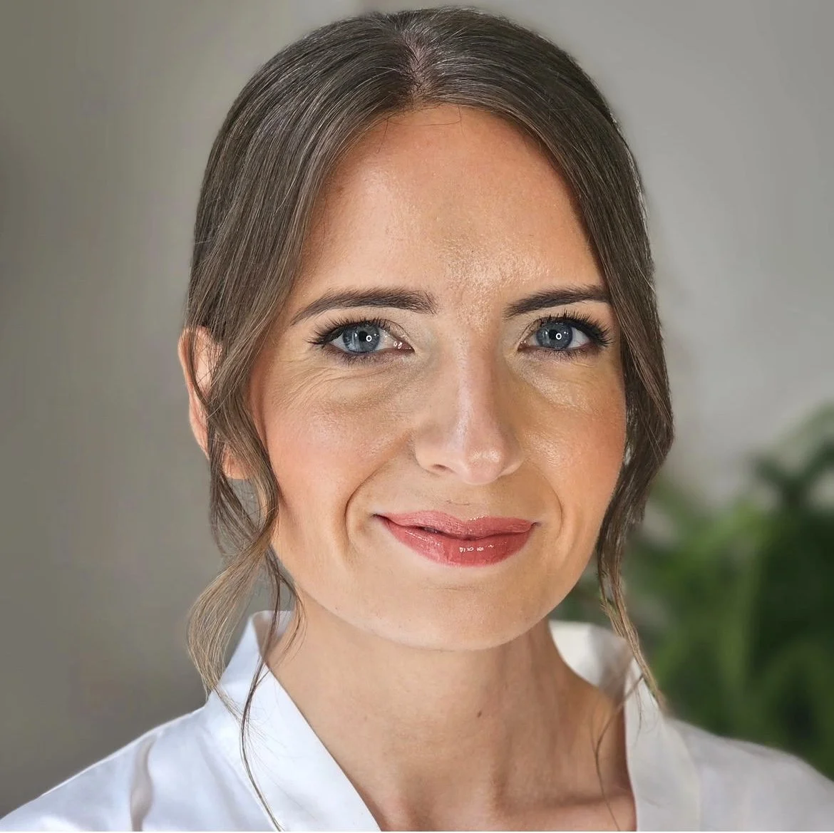 Close-up of a smiling woman with brown hair and blue eyes, wearing a white shirt, with a blurred background.