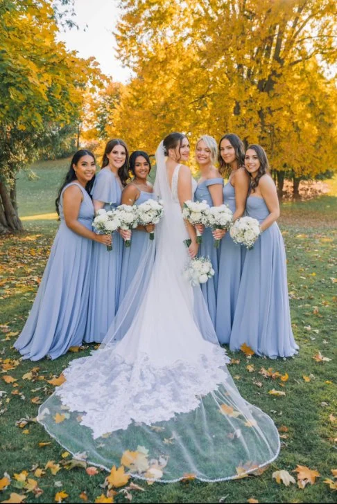 Bride in a white wedding dress and veil standing with bridesmaids in blue dresses, holding bouquets, on a grassy area with autumn foliage.