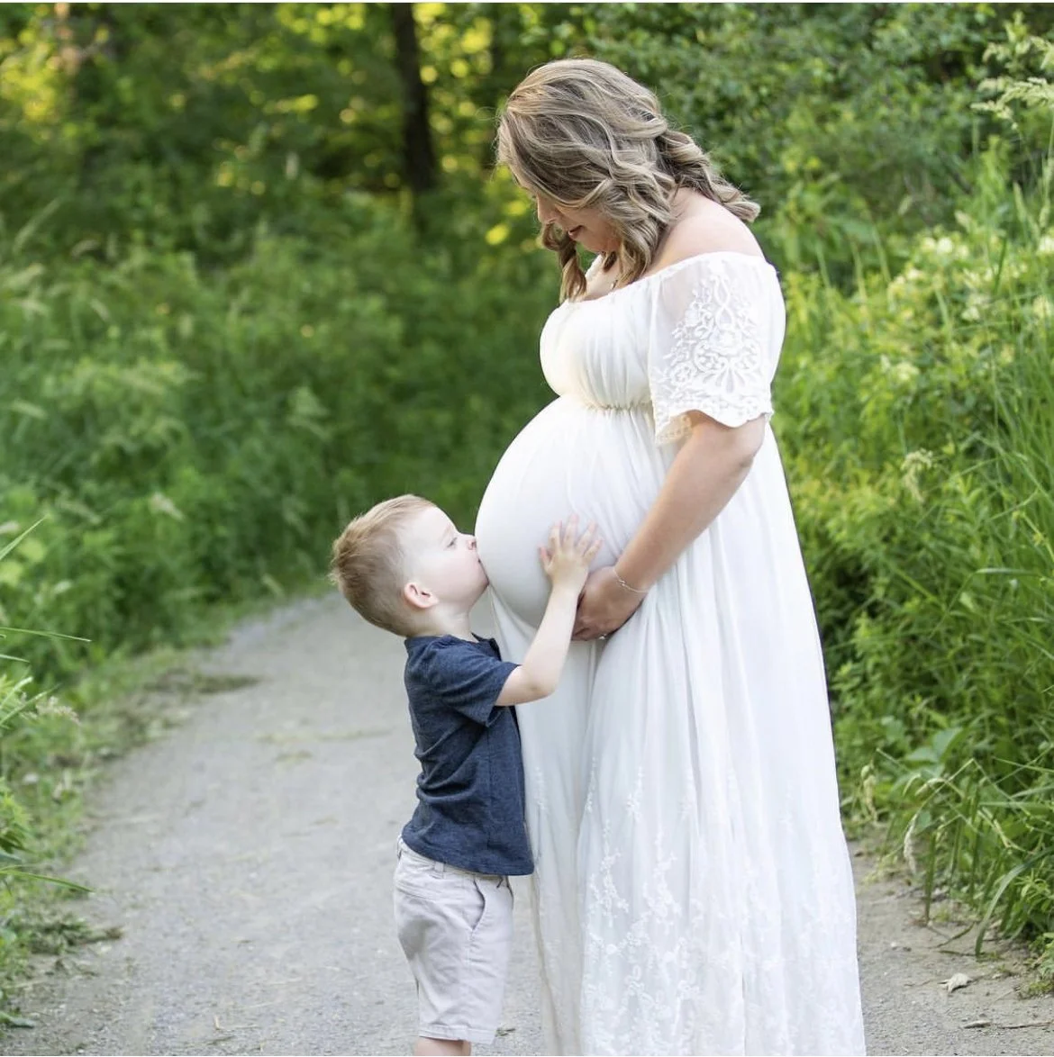 Pregnant woman in white dress with child kissing her belly on a path in a green, wooded area.
