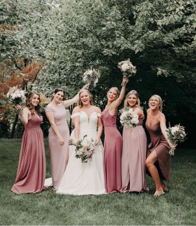 Bride and bridesmaids in pink dresses holding bouquets, posing outdoors on grass with trees in background.