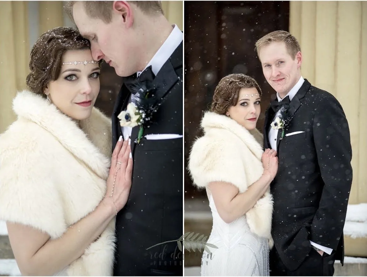 A bride and groom in formal attire stand closely together in a snowy outdoor setting. The bride wears a fur shawl and the groom is in a tuxedo with a boutonniere. Snowflakes fall around them, and they appear affectionate and happy.
