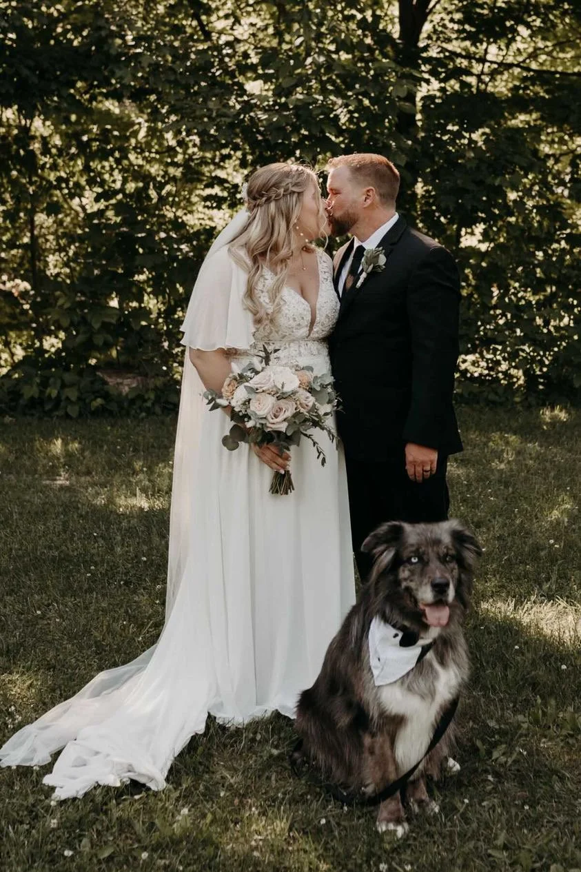 Bride and groom kissing, holding a bouquet, with a dog in a tuxedo bandana, outdoors.