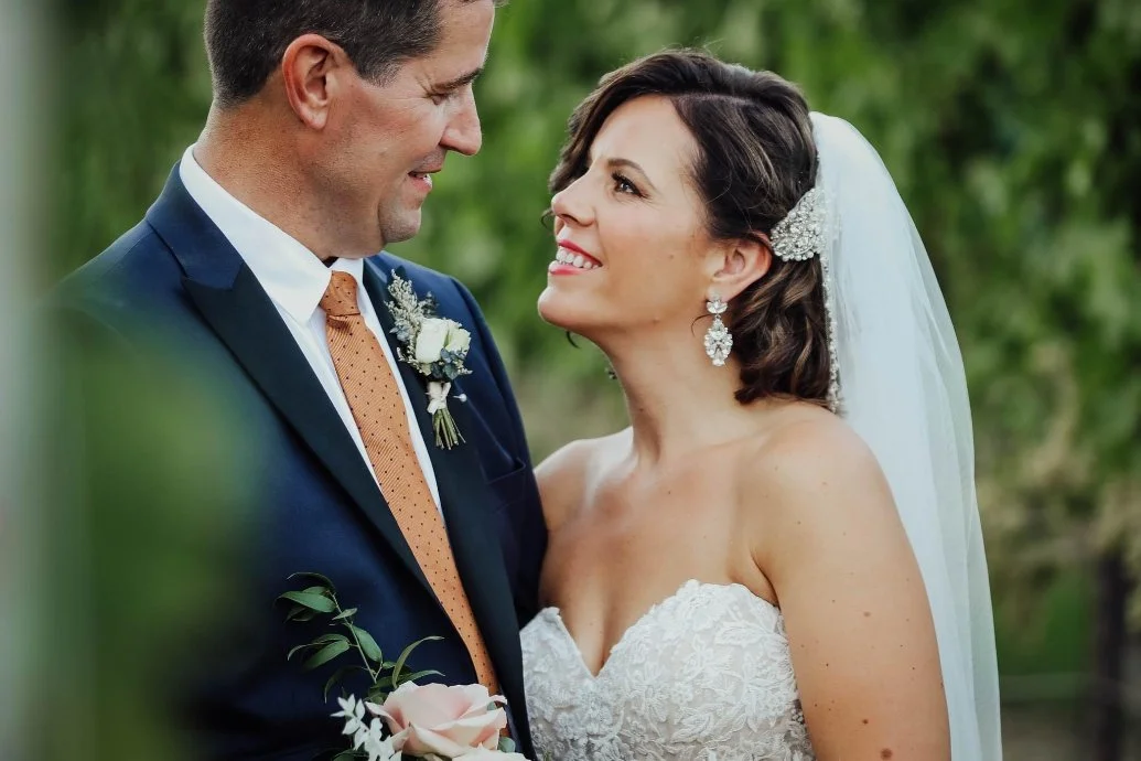 Bride and groom smiling at each other outdoors, with the groom in a navy suit holding a bouquet and the bride wearing a lace wedding gown and veil.