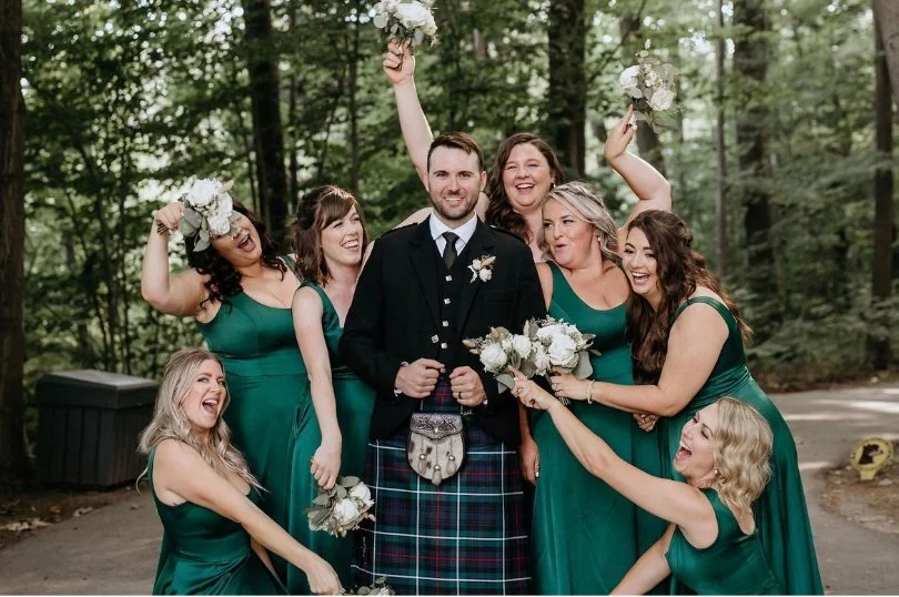 Groom in traditional Scottish kilt surrounded by joyful bridesmaids in green dresses, holding bouquets, against a forest backdrop.