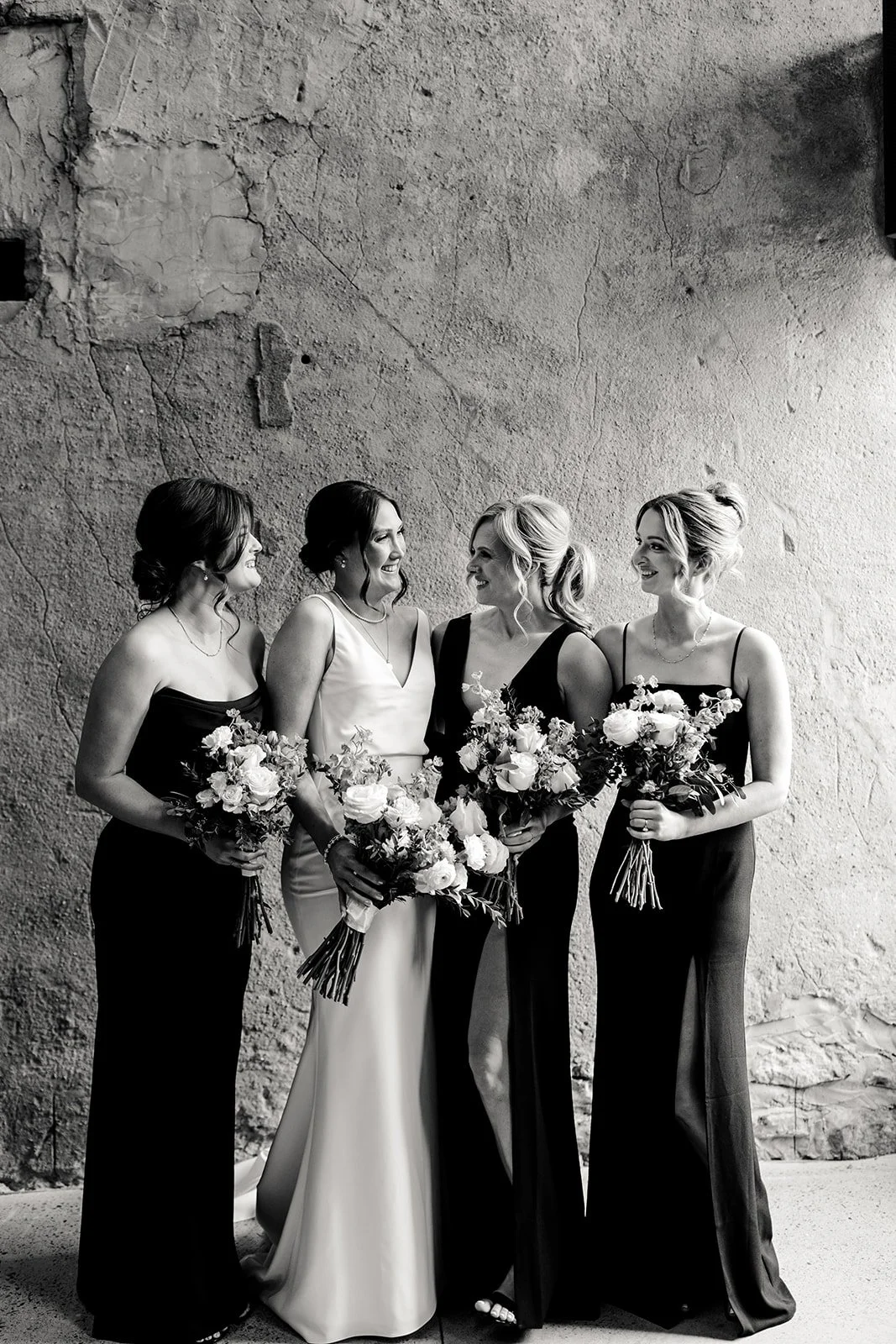 Black and white photo of four women in formal dresses holding bouquets, standing in front of a textured wall, smiling and looking at each other.