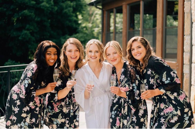 A bride and four bridesmaids in floral robes celebrating with champagne outdoors.