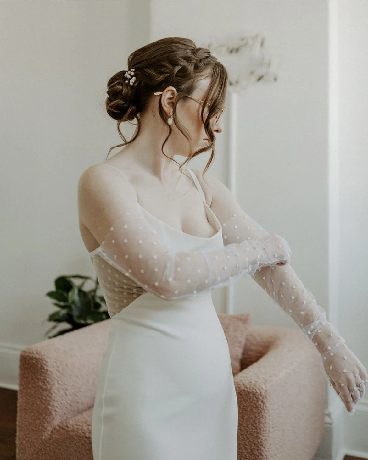Woman in a wedding dress adjusting sheer sleeves in a softly lit room.