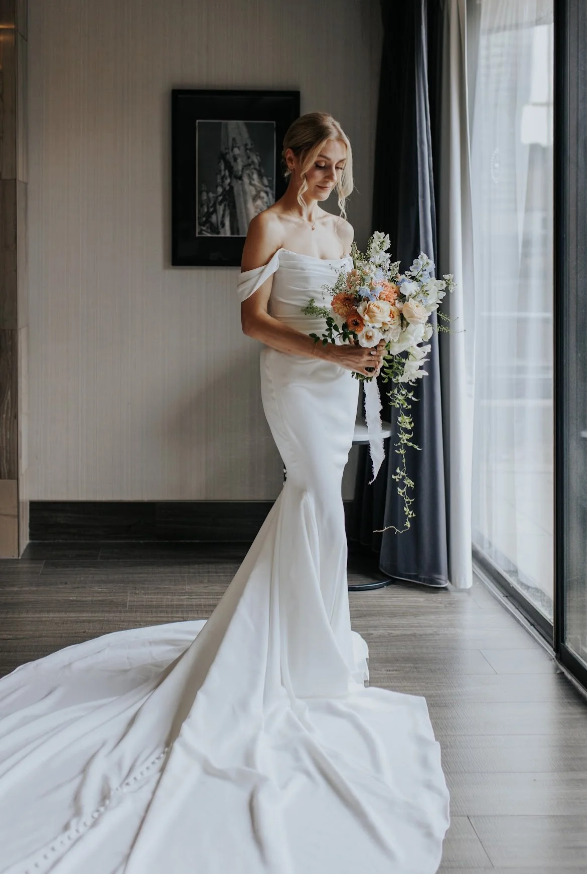 Bride in an off-the-shoulder white wedding dress holding a bouquet, standing indoors by a window.