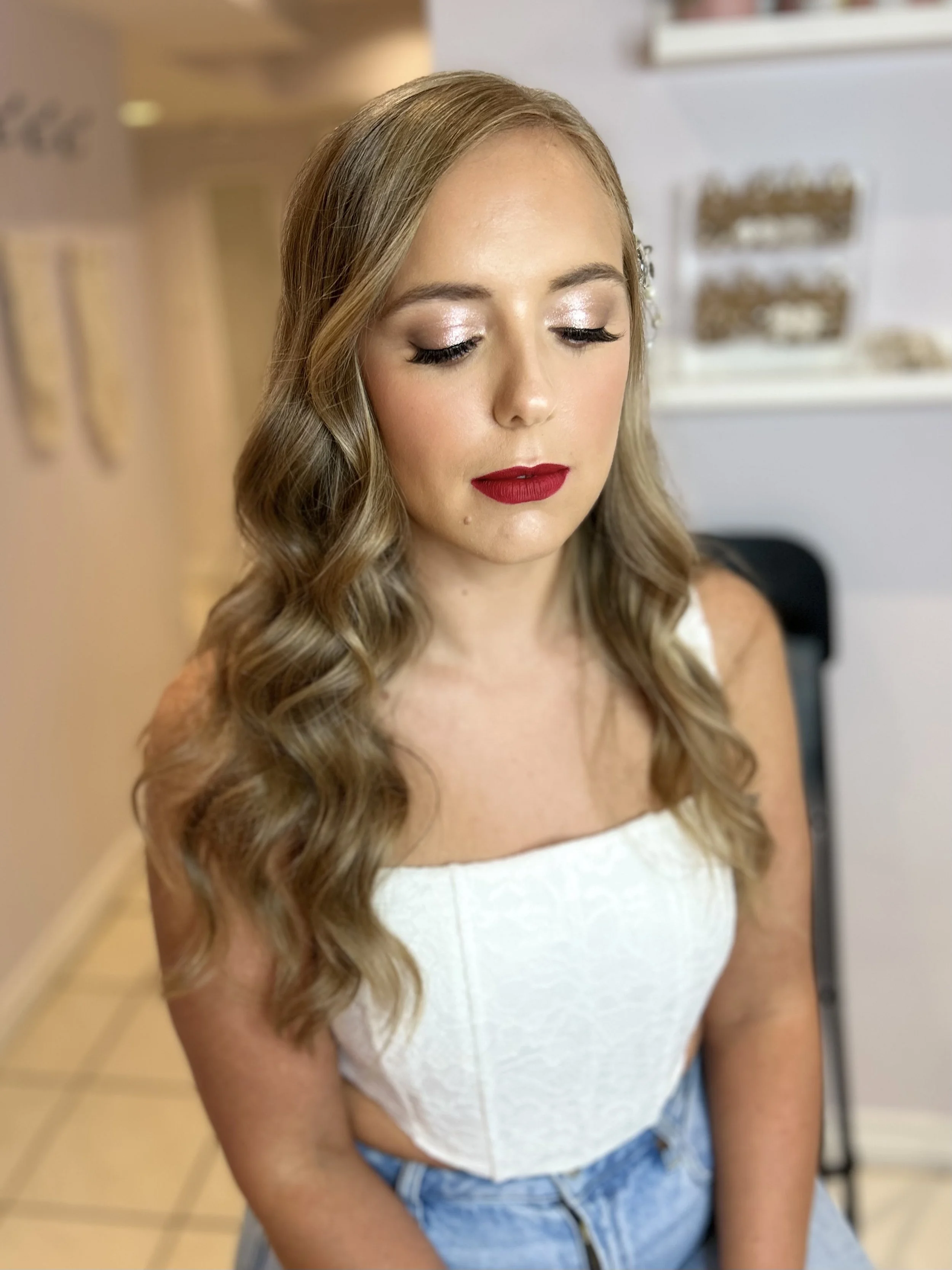 Woman with closed eyes, wearing white top, red lipstick, and wavy hair, in indoor setting.
