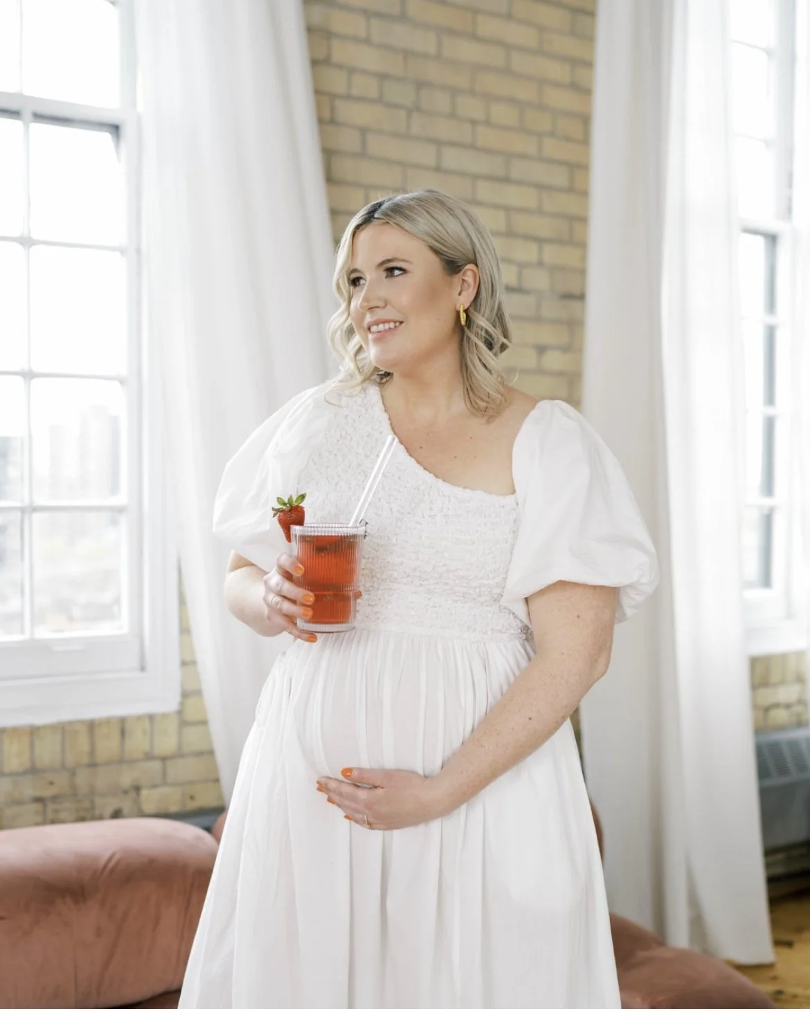 Pregnant woman in white dress holding a drink with a strawberry garnish.