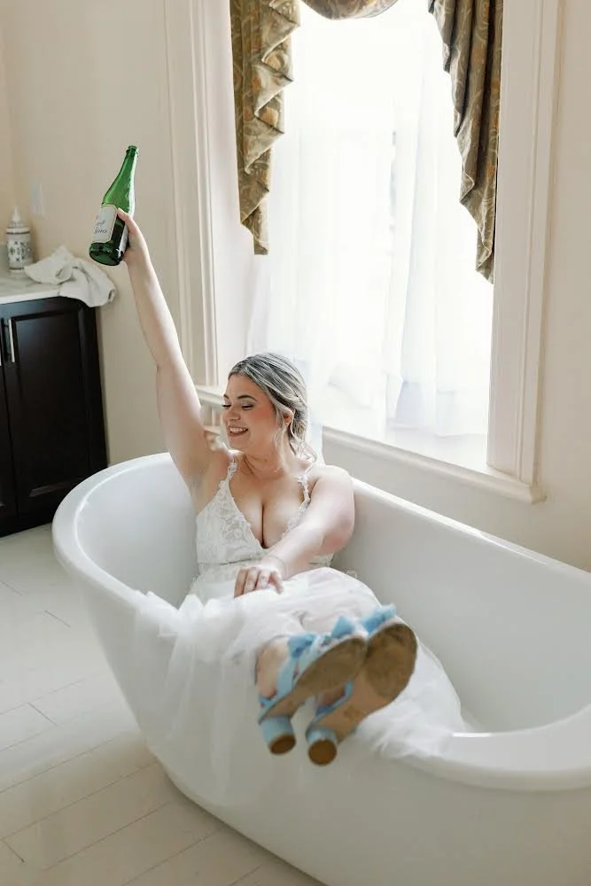 Woman in wedding dress sitting in bathtub holding a champagne bottle.