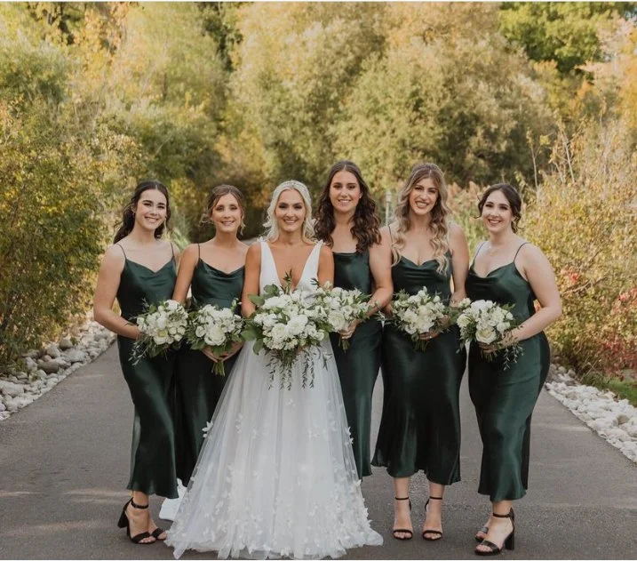 Bride with bridesmaids in green dresses holding bouquets, standing on a pathway surrounded by greenery.