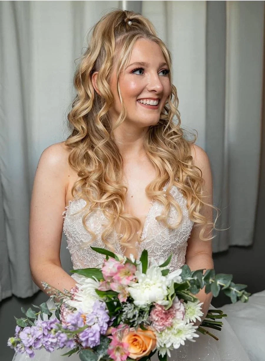 Smiling bride holding a bouquet of flowers, wearing a lace wedding dress with long, curly hair styled in a half-updo.