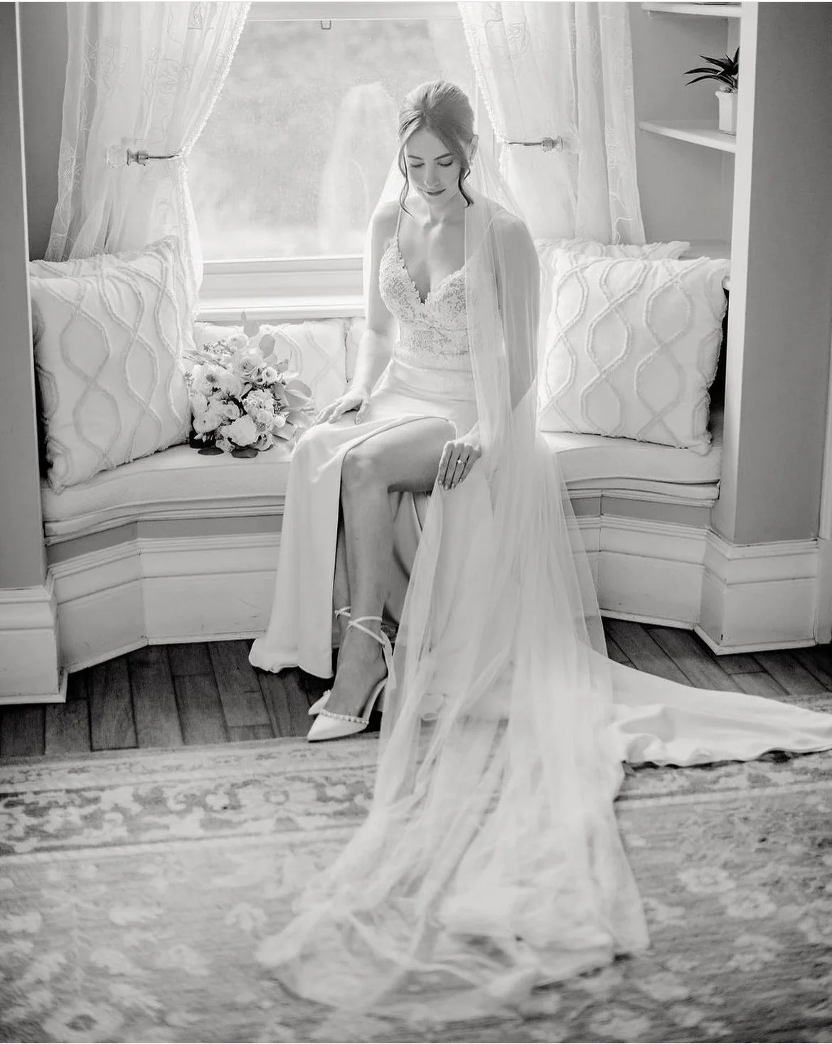 Bride sitting on window seat with long veil and bouquet, black and white photo