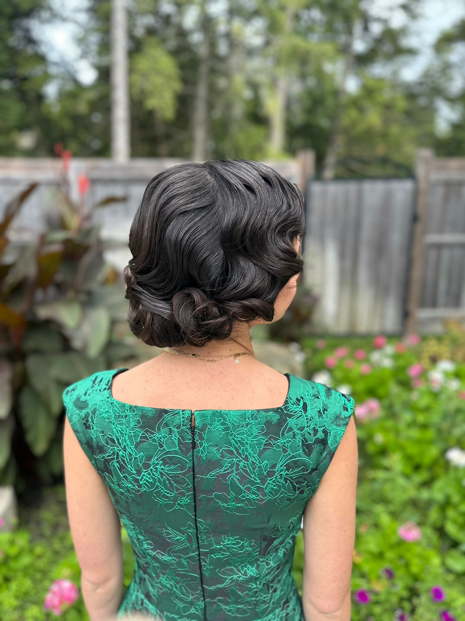 Woman in vintage green dress with finger wave hairstyle standing outdoors with blurred garden background.
